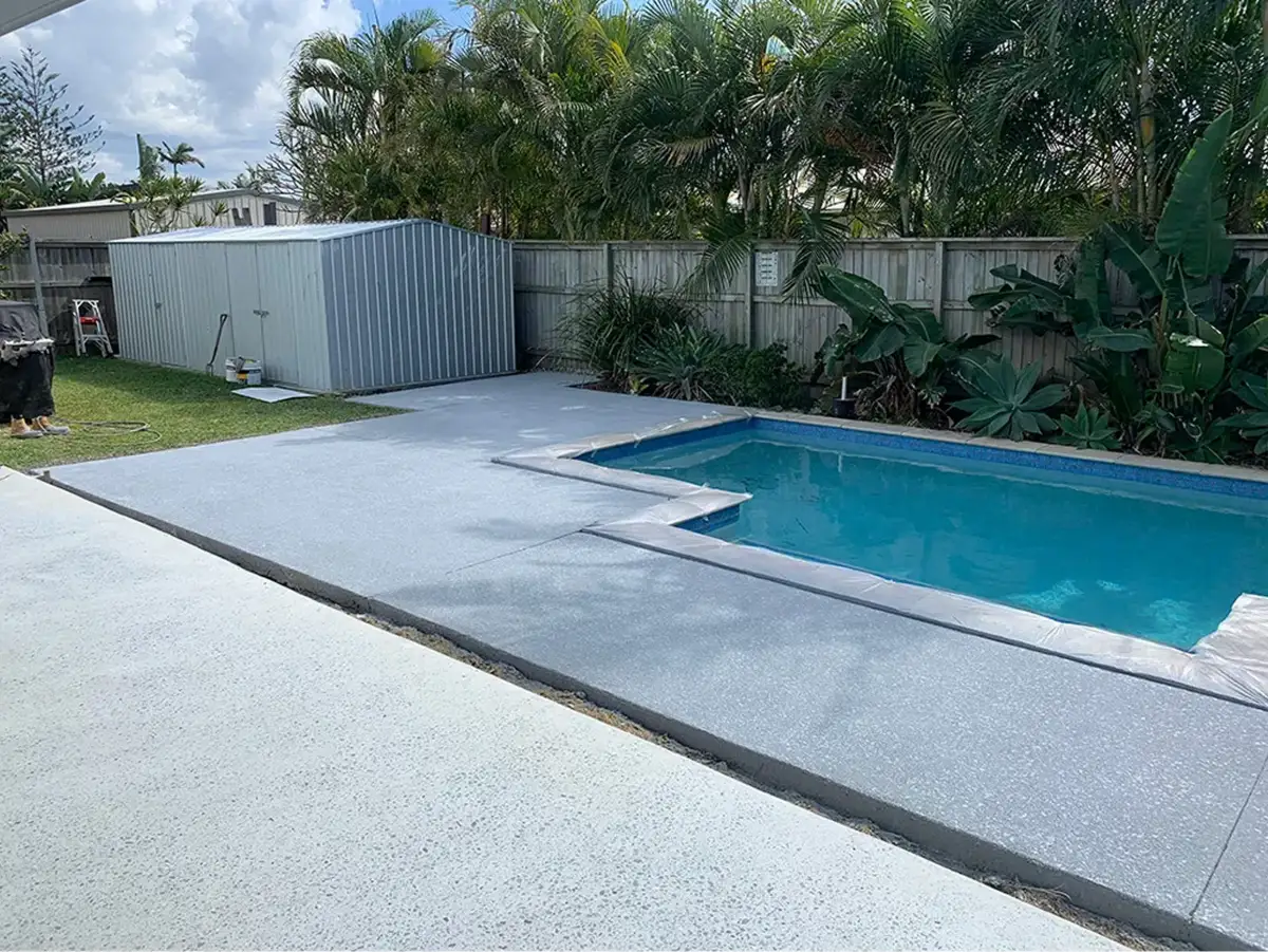 Light grey resurfaced concrete pool surround beside a swimming pool, shed and palm trees
