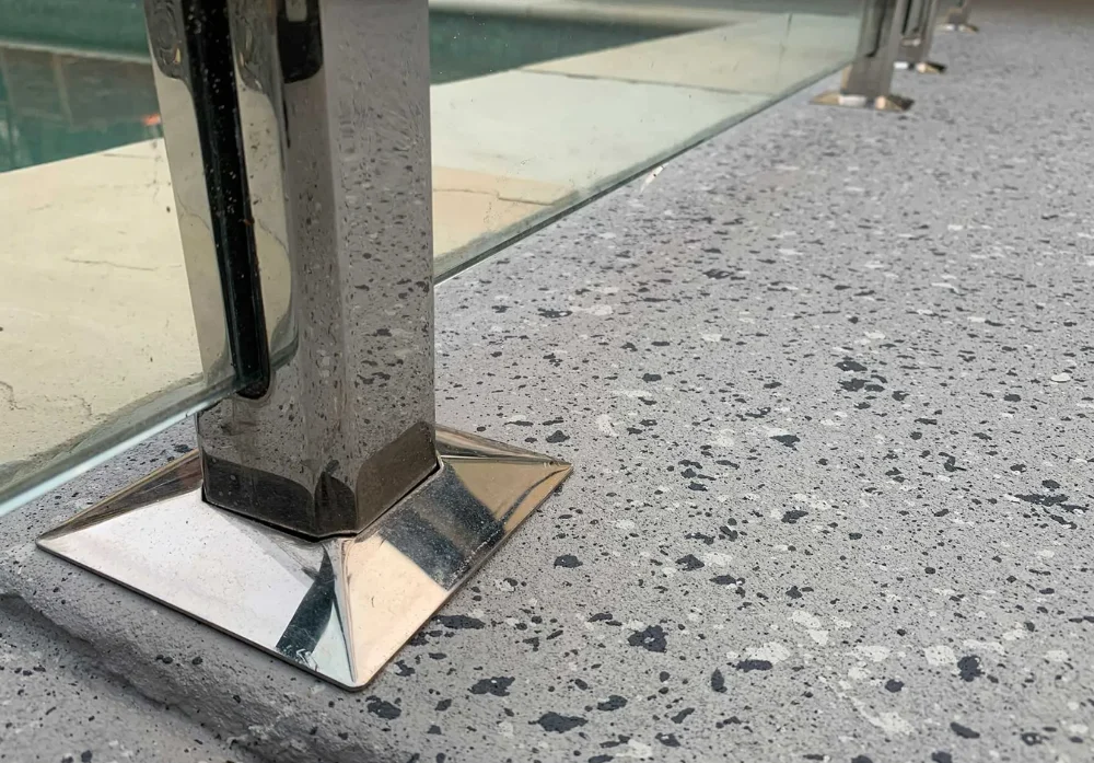 Close-up of speckled exposed aggregate concrete beside a steel post and glass pool fence