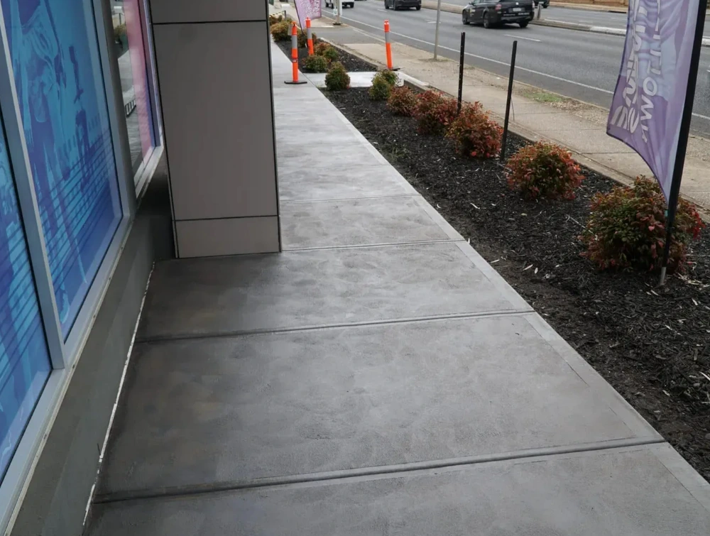 Grey concrete footpath beside a garden bed and a building, next to a road