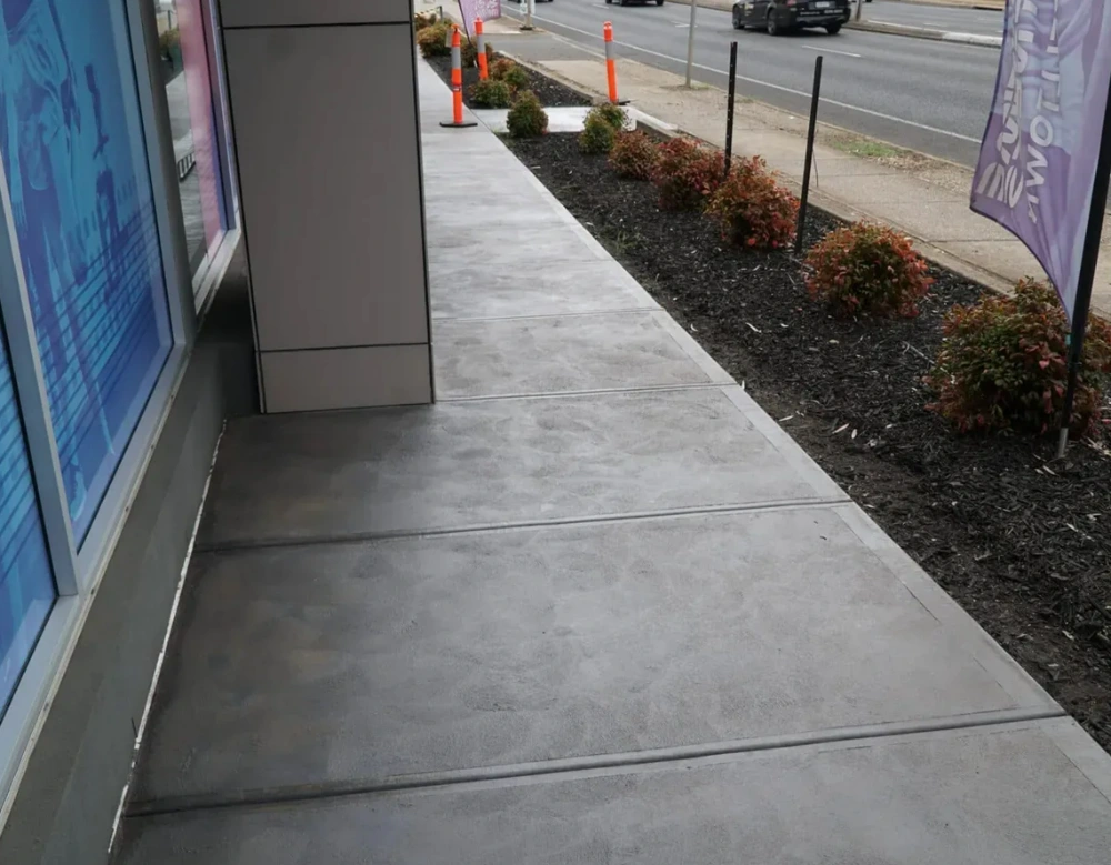 Grey concrete footpath beside a garden bed and a building entrance next to a road