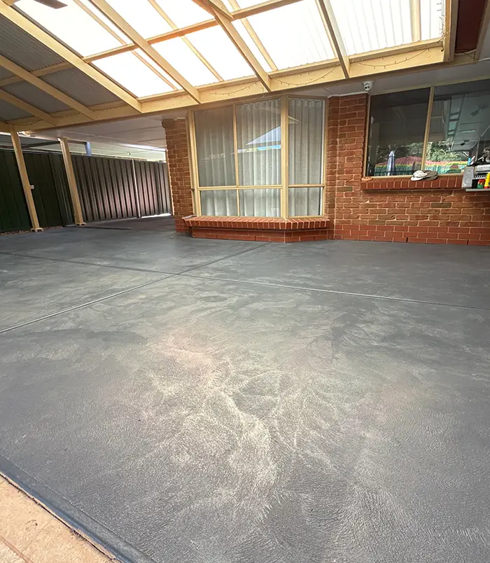 Rough concrete patio floor under a pergola beside a brick wall, before epoxy flooring