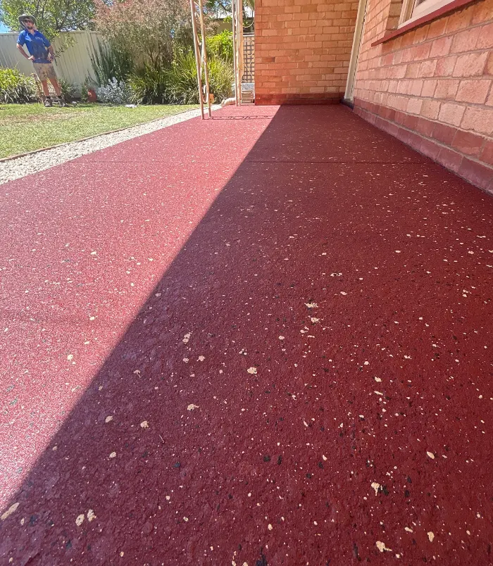 Deep red resurfaced concrete path with a speckled finish beside a brick wall and lawn