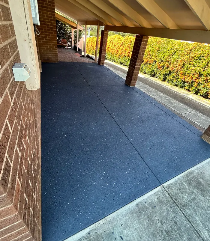 Dark resurfaced concrete floor under a carport beside a brick wall and orange hedge