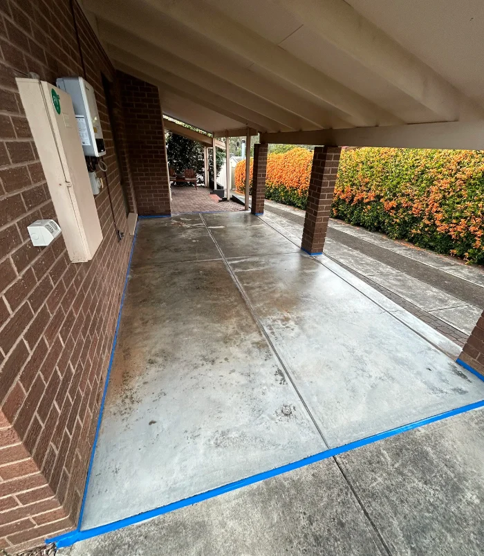Stained, patchy concrete floor under a carport beside a brick wall and orange hedge, before resurfacing