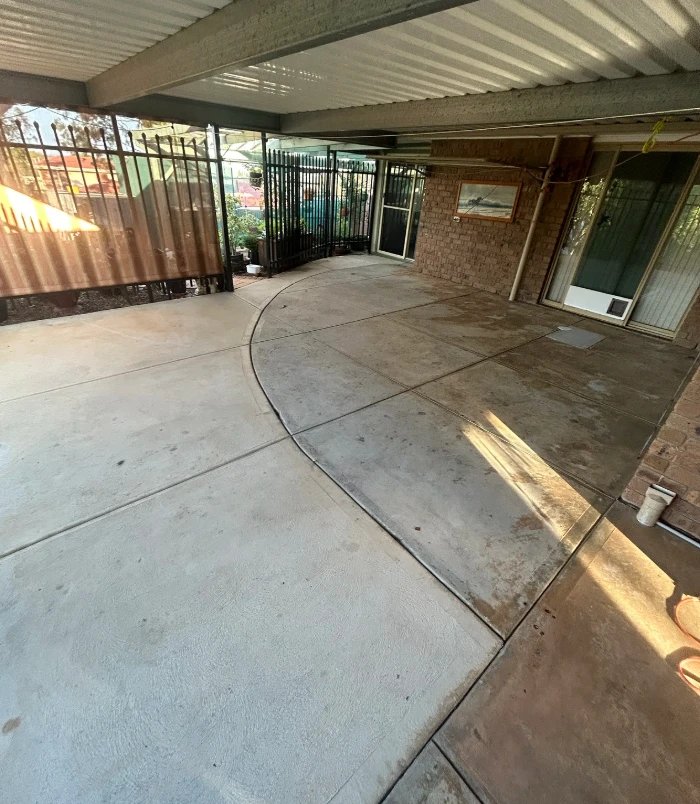 Worn, stained concrete patio under a pergola beside a black fence and glass door, before resurfacing