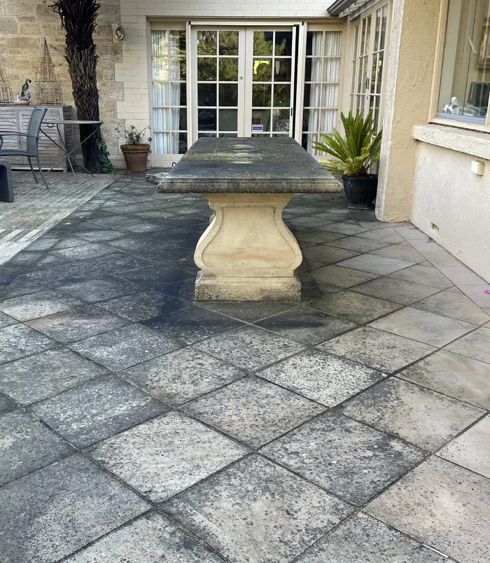 Dark, stained pavers around a stone outdoor table beside French doors, before paver cleaning