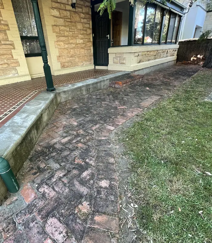 Dirty, algae-stained red brick paving beside a sandstone house, before paver cleaning