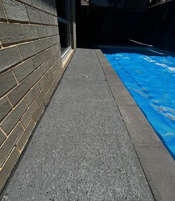 Speckled grey resurfaced pool surround beside a dark brick wall and pool cover