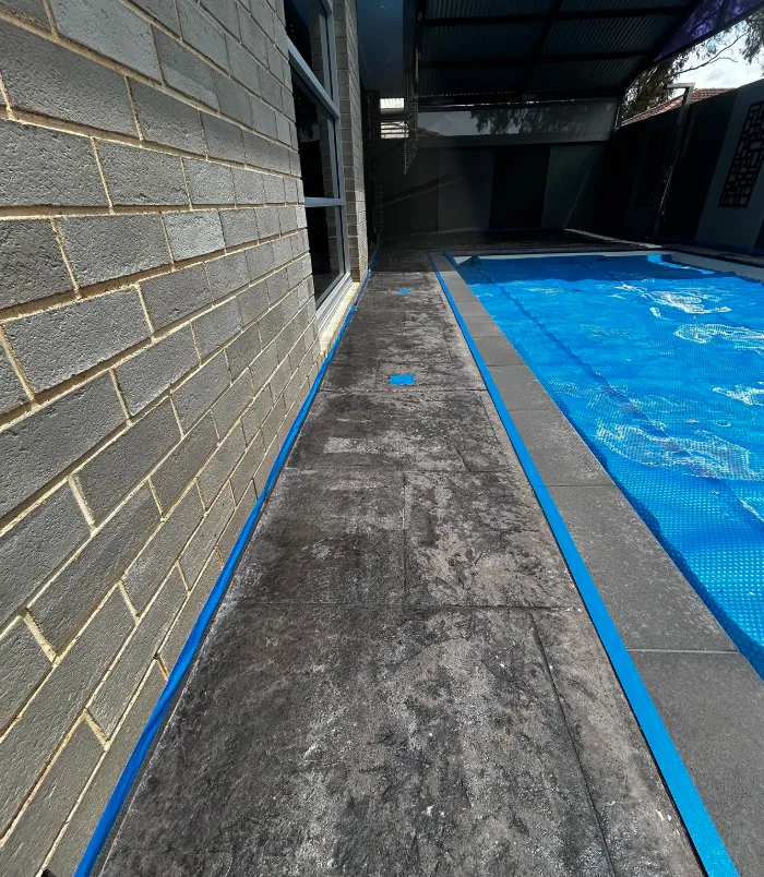 Stained concrete pool surround with blue masking tape beside a dark brick wall, before resurfacing