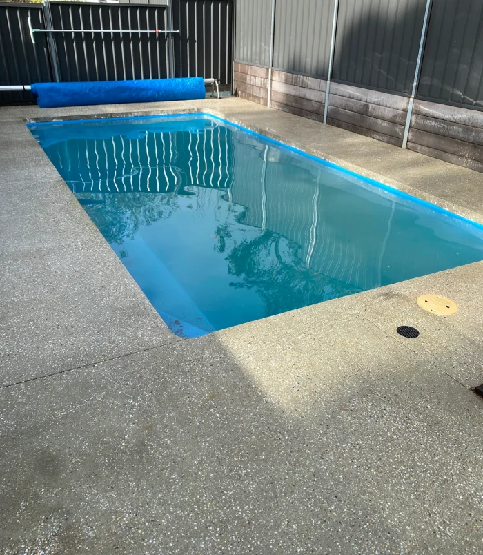 Plain concrete pool surround beside a pool with a rolled-up cover, before pool resurfacing