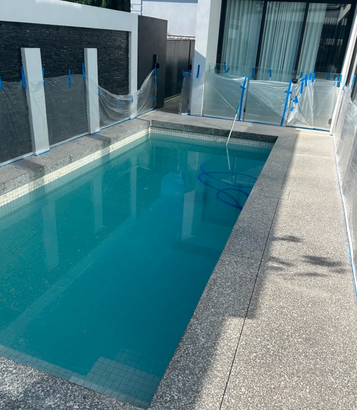 Concrete pool surround with blue masking tape beside a glass pool fence, before pool resurfacing
