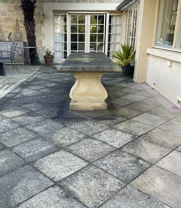 Dark, stained pavers around a stone outdoor table beside French doors, before pressure washing