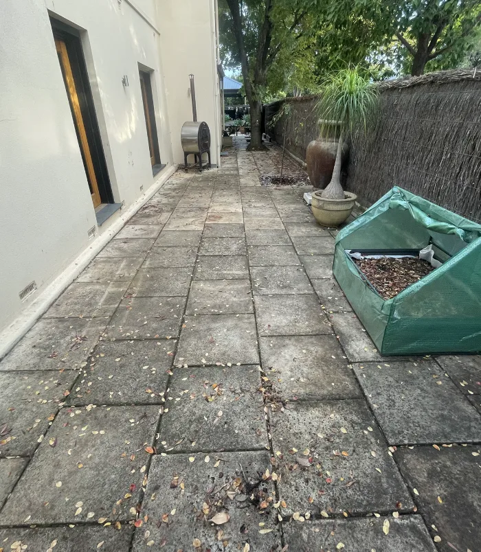 Dark, stained pavers in a side courtyard beside a rendered wall and garden bed, before pressure washing